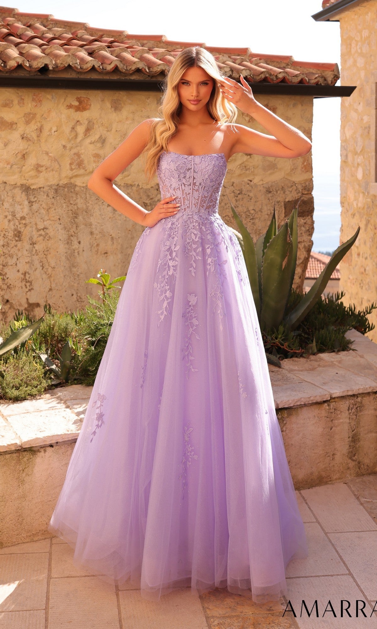A woman poses gracefully outdoors in front of a stone wall and lush plants, wearing the Amarra 88950 Strapless Embroidered Prom Ball Gown with a sweetheart neckline and fitted bodice.